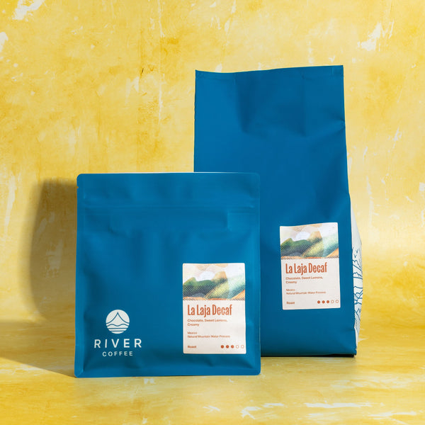 Two blue coffee bags labeled 'La Laja Decaf' by 'River Coffee' on a yellow textured background.