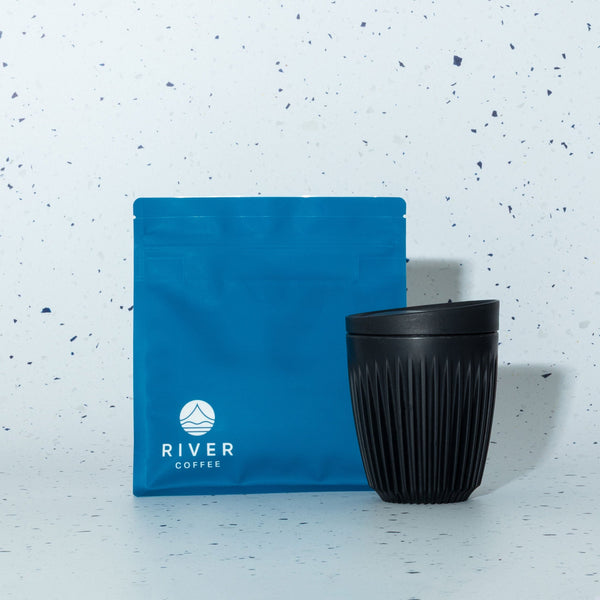 Black coffee cup and blue coffee bag with 'River Coffee' logo on a light speckled background