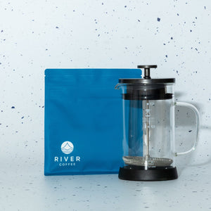 Clear French press coffee maker with black lid and base, next to a blue River Coffee bag on a light blue speckled background.