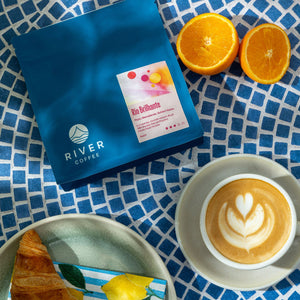 Blue coffee bag with 'Rio Brillante' label, orange slices, and a cup of coffee with latte art on a blue and white checkered tablecloth.