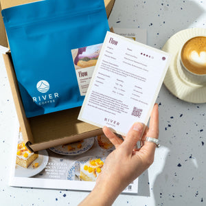 Person holding a card with 'River Coffee' branding next to a coffee cup and package on a speckled surface.