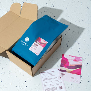 Open cardboard box with a blue coffee bag labeled 'River Coffee' on a speckled surface.