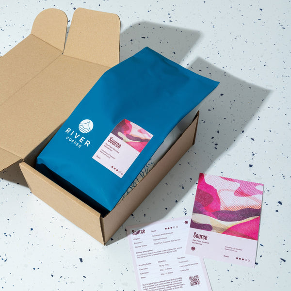 Open cardboard box with a blue coffee bag labeled 'River Coffee' on a speckled surface.