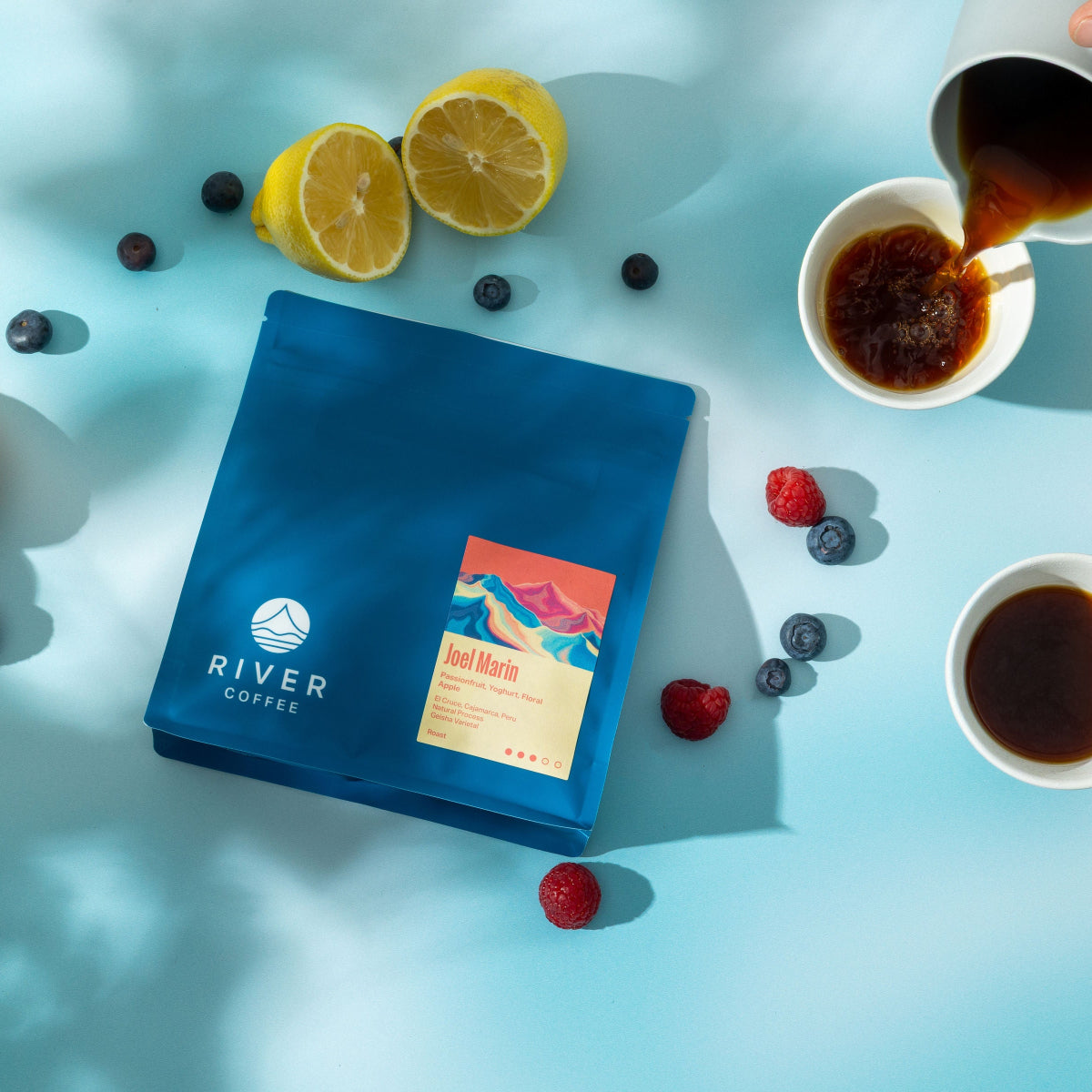 A blue bag of River Coffee Roasters coffee is displayed in a styled flat-lay on a light blue surface, surrounded by citrus fruits, berries, and cups of freshly brewed coffee, with a hand pouring coffee in the top right corner.